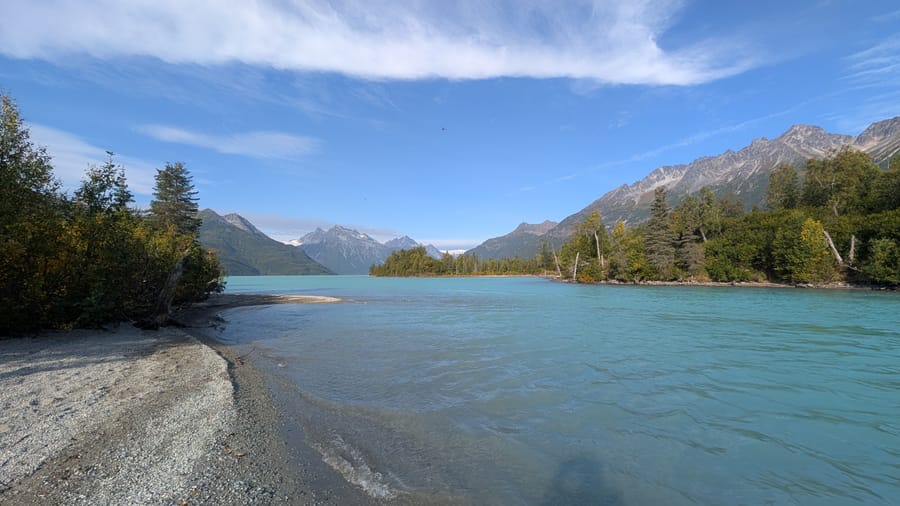 From the southern shore of Crescent Lake, southwest of Mt. Redoubt, looking west. Aquamarine glacial-colored waters lap the shore under a blue sky with wispy clouds.
